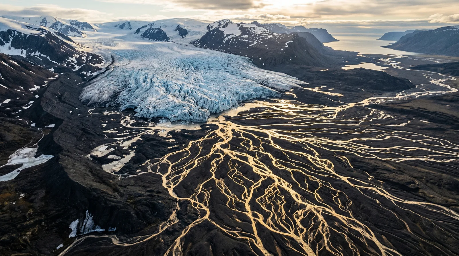 Aerial view of a glacier feeding golden braided rivers across a dark valley at sunset.