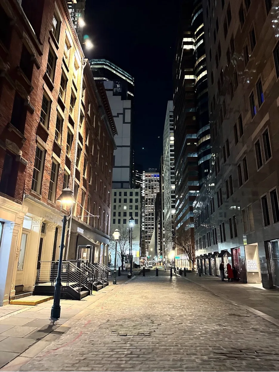 Empty cobblestone city street at night flanked by tall buildings and warm lit windows.