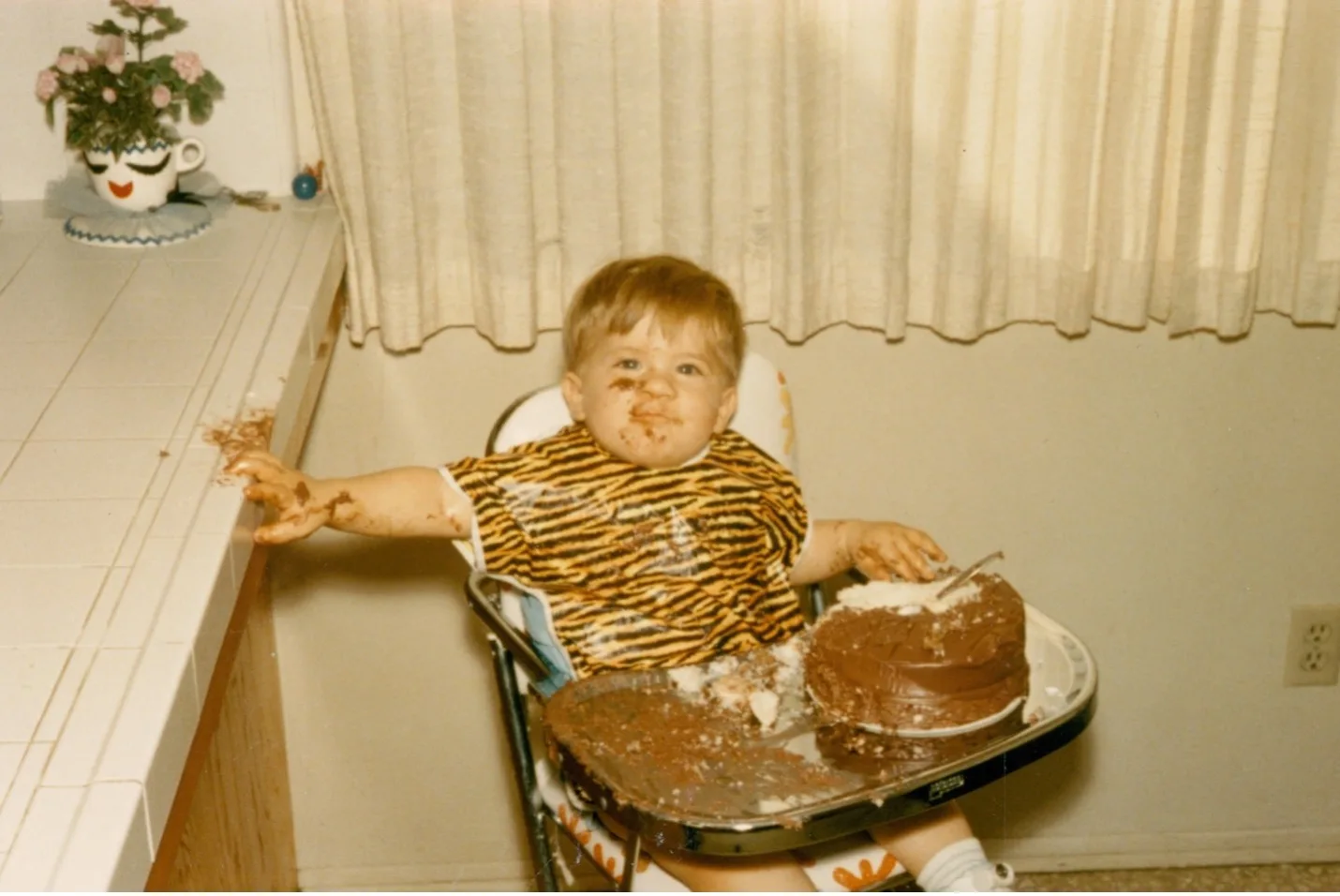Vintage photo of a toddler in a tiger shirt in a high chair, face and hands covered in chocolate cake.