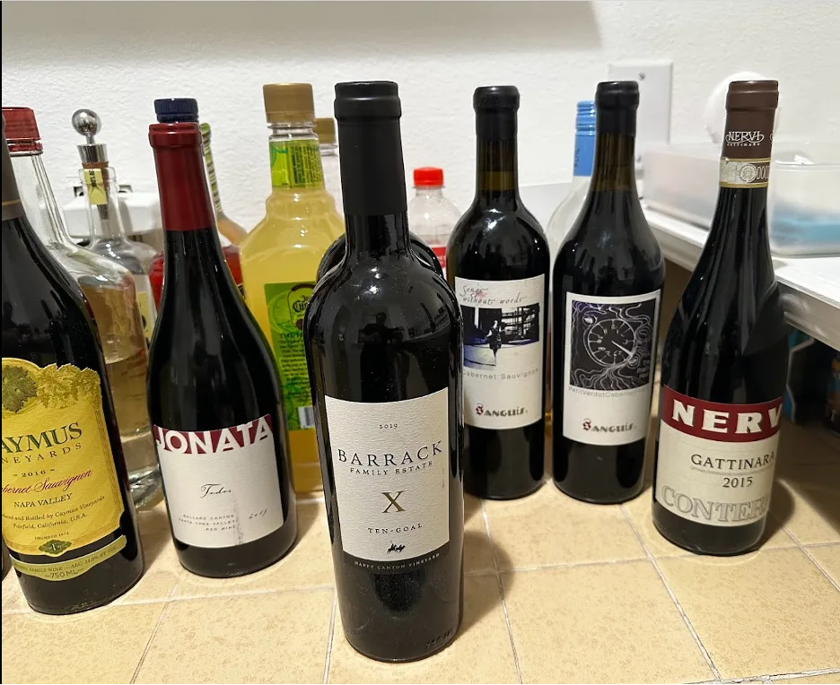 Row of red wine bottles on a tiled counter, including Caymus, Jonata, Barrack Family Estate, and Nervi Gattinara.