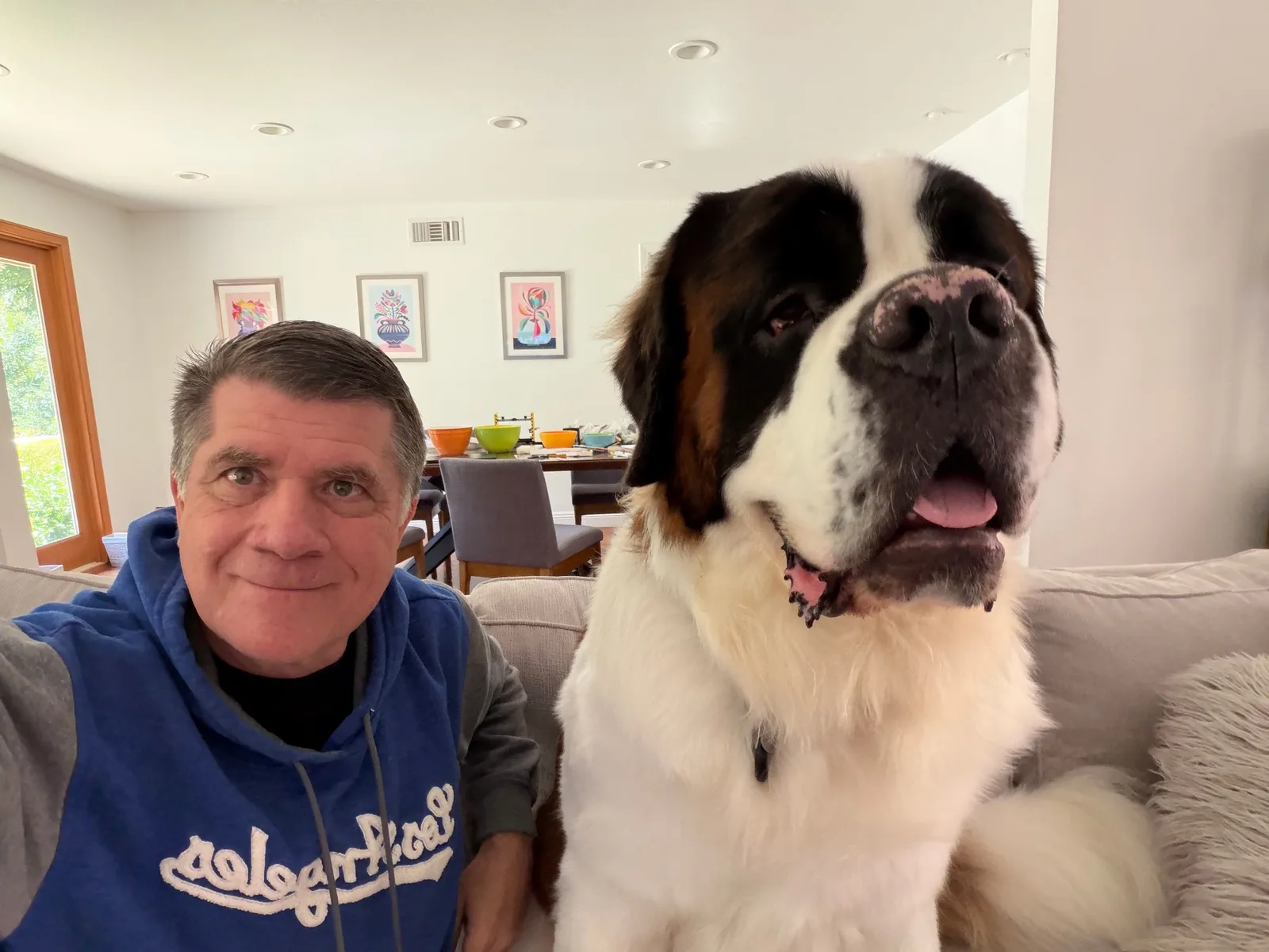 Steve Smith takes a selfie on a living room couch beside Magnus, his Saint Bernard, who sits upright next to him with his head raised and tongue out, nearly as tall as Steve is sitting.