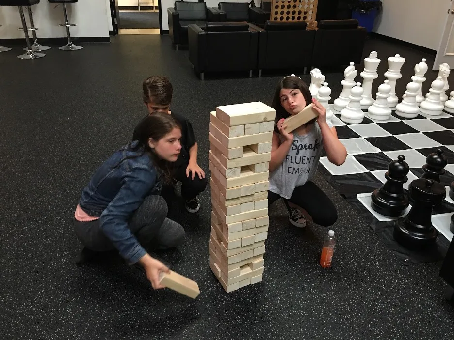 Three young people playing a giant Jenga block tower near an oversized floor chess set