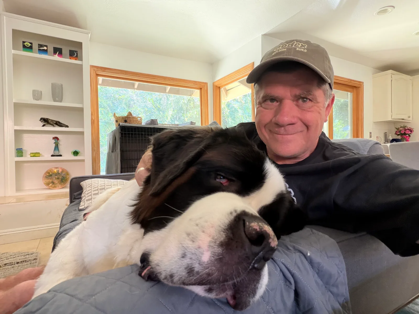 Steve Smith in a ball cap takes a selfie on the couch with Magnus, his Saint Bernard, stretched out across his lap with his head resting on a blanket.