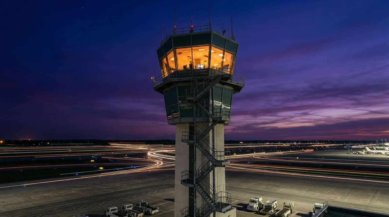 Lit air traffic control tower at dusk above runways streaked with light trails.