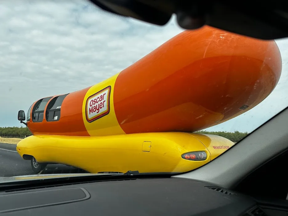 The Oscar Mayer Wienermobile, a giant hot dog shaped vehicle, seen through a car windshield