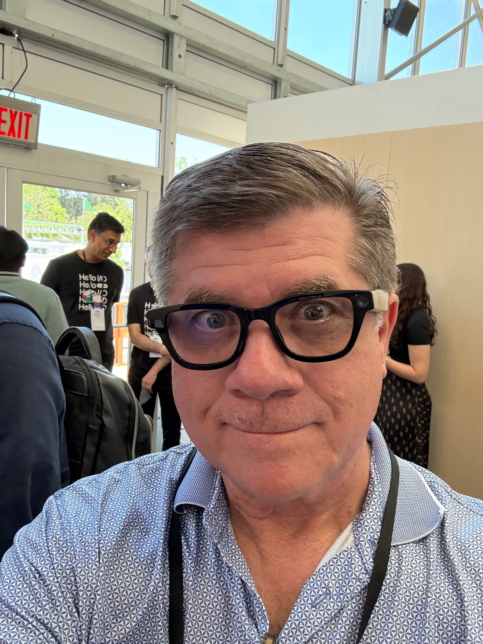 Man in dark glasses takes a selfie near an exit door at a crowded conference venue