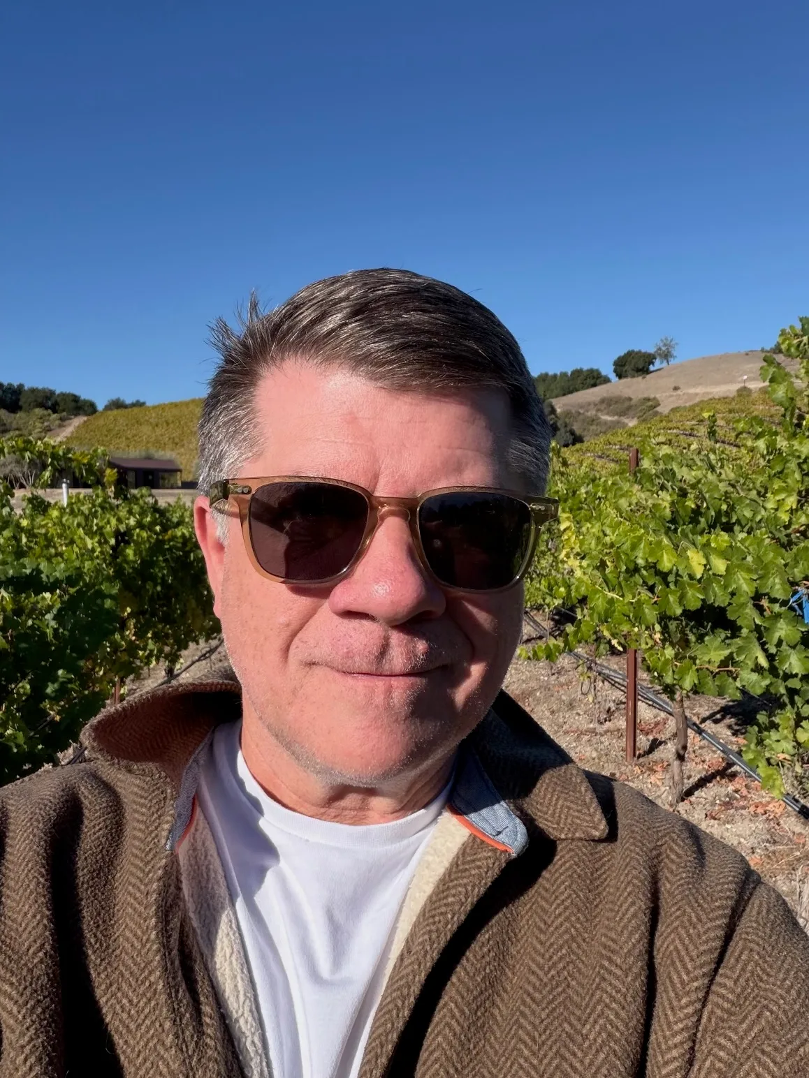 Steve Smith in sunglasses and a brown herringbone jacket takes a selfie among the vineyards in Paso Robles, with vine rows and a golden hillside behind him.
